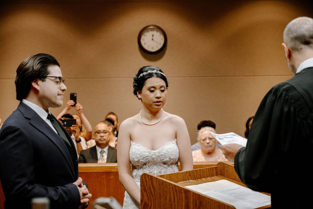 A wedding ceremony takes place in a courtroom with participants facing a judge at the bench while exchanging vows.