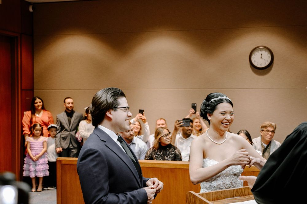 People laugh during an indoor ceremony in a courtroom or municipal building with warm lighting and a clock on the wall.