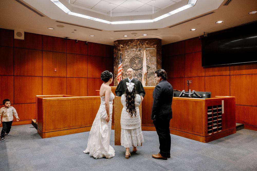 A wedding ceremony takes place in a courtroom with warm wood paneling and an octagonal ceiling design.
