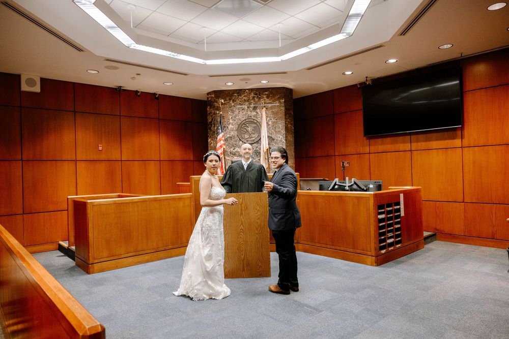 Wedding ceremony takes place in a wood-paneled courtroom with domed ceiling and judge's bench.