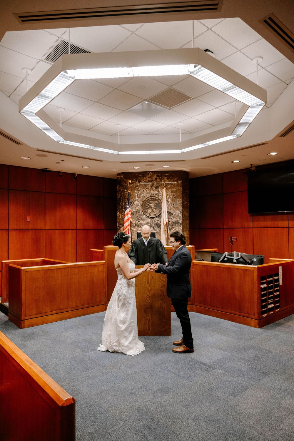 A wedding ceremony taking place in a courtroom with an octagonal ceiling light fixture and wooden paneled walls.