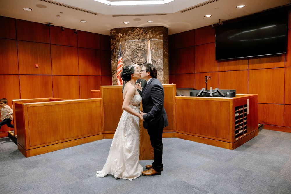 Two people share their first kiss as newlyweds in front of a wood-paneled courtroom bench.