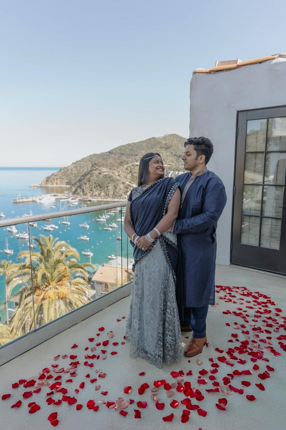 Couple stands on balcony overlooking harbor with red rose petals scattered at their feet.