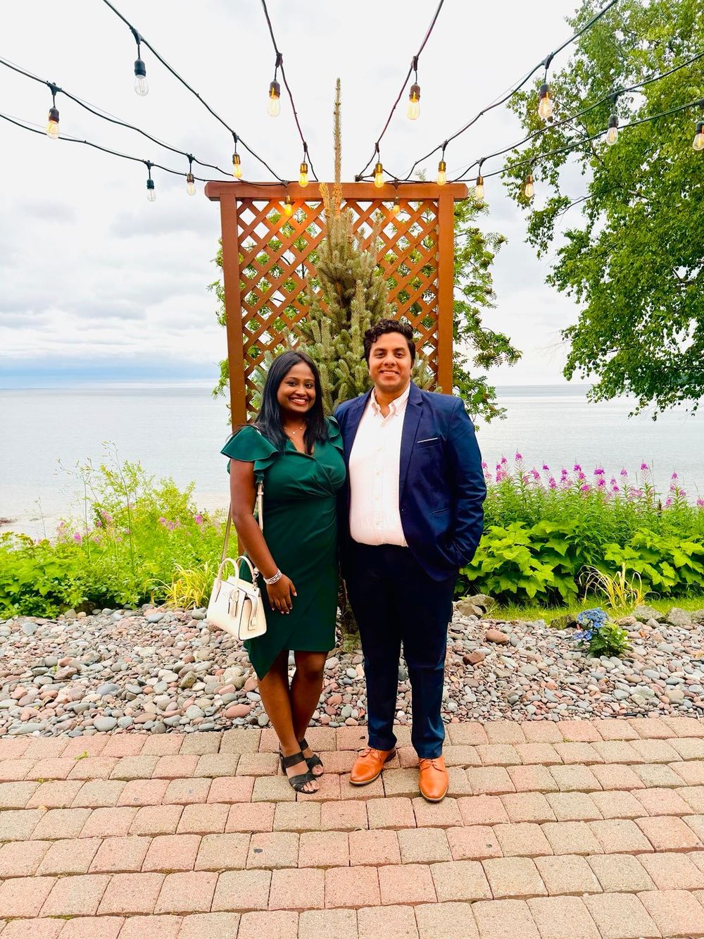 Couple posing in front of wooden pergola with string lights and waterfront view.