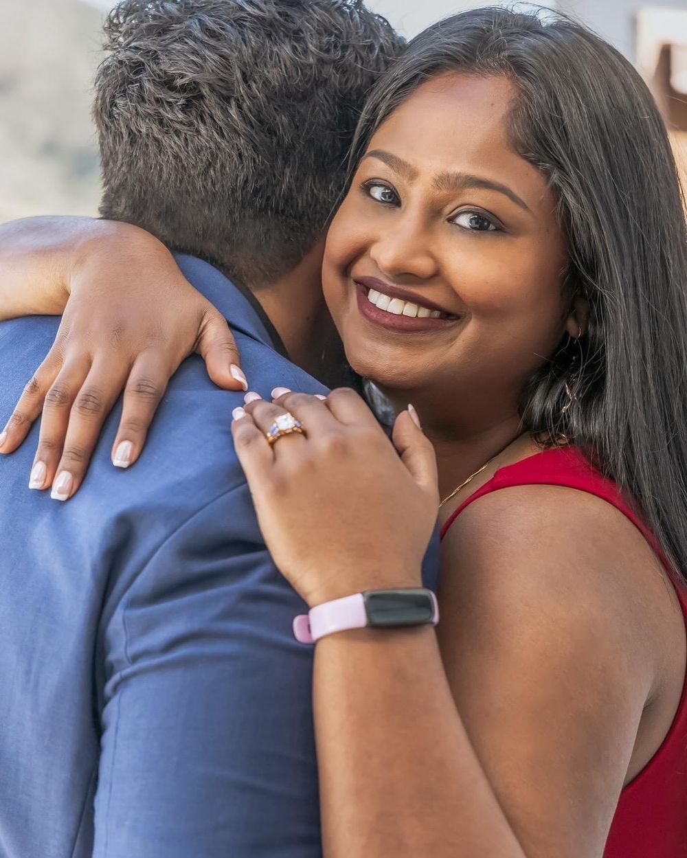 A couple sharing a loving embrace with a red dress and blue suit clothing visible, showing engagement ring.
