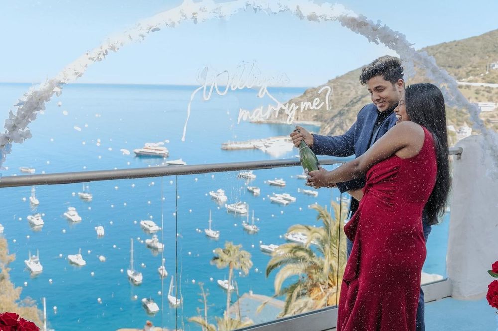 A couple enjoys a scenic overlook of the Mediterranean Sea from a white arched terrace with boats visible in the harbor below.