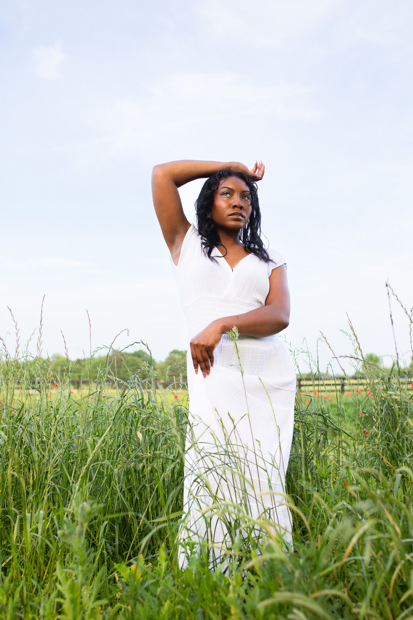 A woman in a flowing white outfit stands gracefully among tall grass in a serene outdoor field setting.