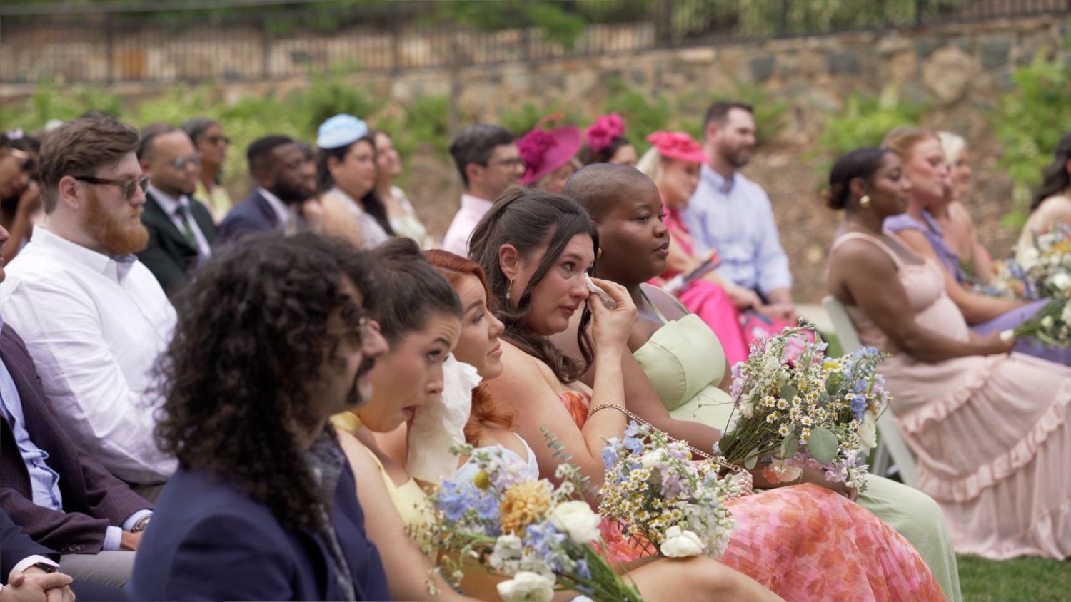 A row of wedding guests seated outdoors at a ceremony with flower bouquets and pastel-colored attire.