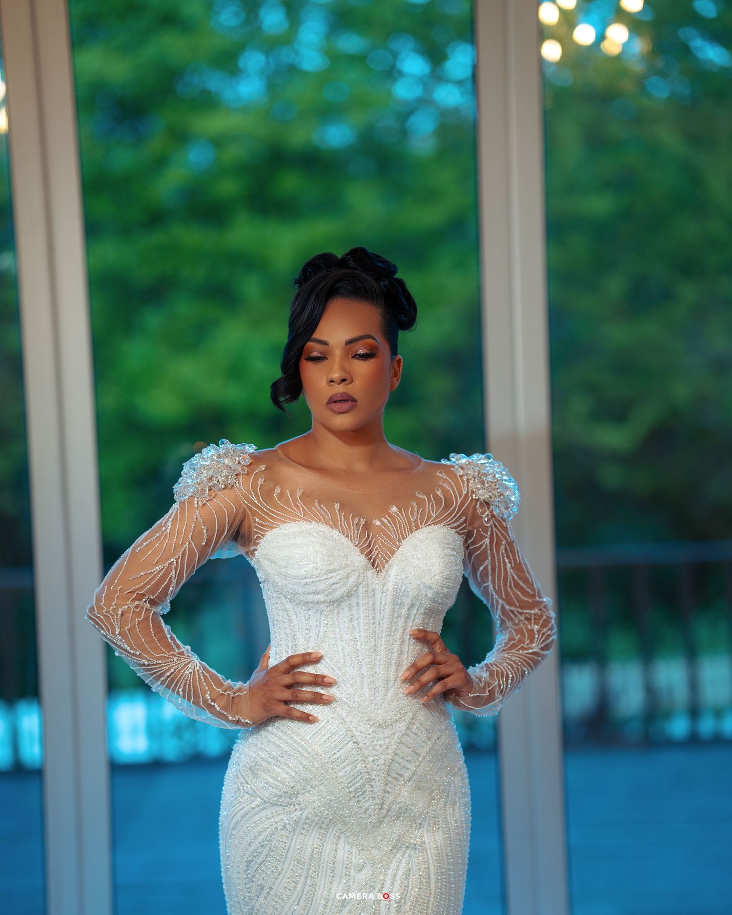 An elegant bridal portrait showing a fitted white wedding gown with sheer lace sleeves and intricate beading in a modern setting.