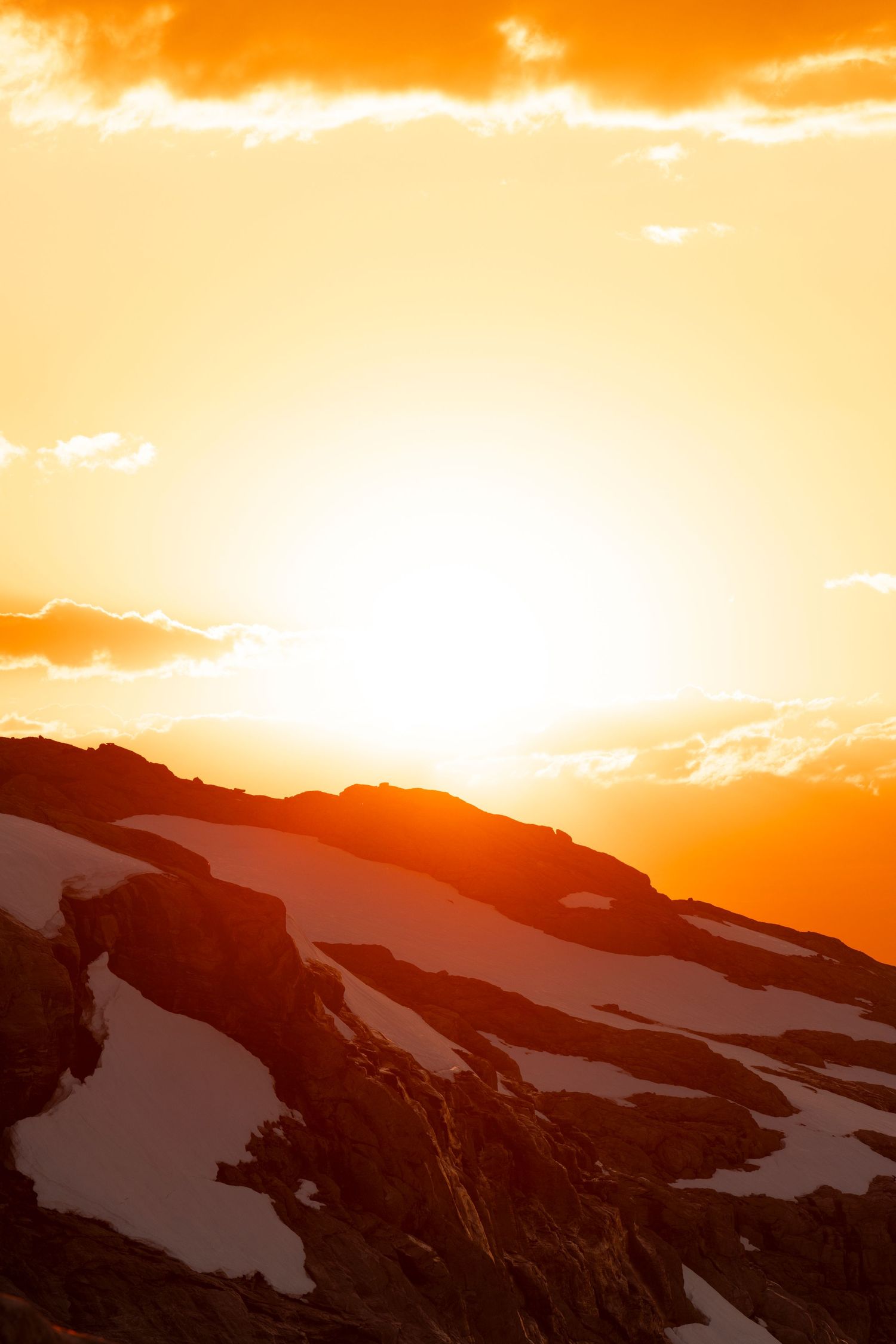 Dramatic orange sunset illuminates snowy mountain peaks with a bright glowing sky and wispy clouds above.