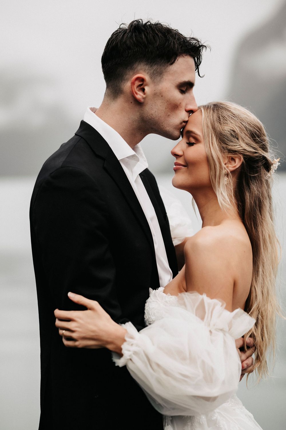 A romantic wedding couple embraces in an intimate moment against a moody cloudy backdrop.