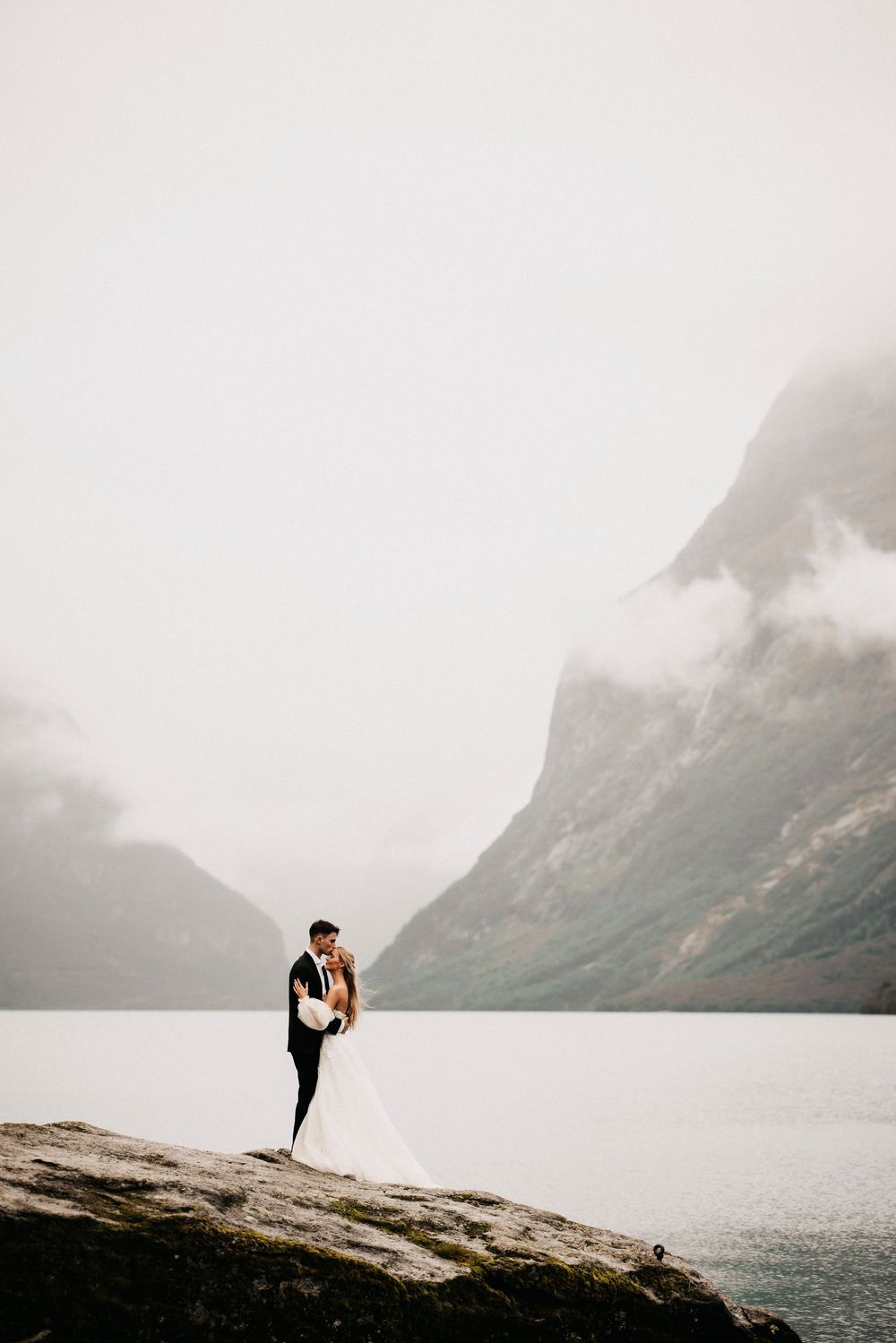 A couple in wedding attire embracing on a rocky outcrop overlooking a misty mountain lake in dramatic landscape.