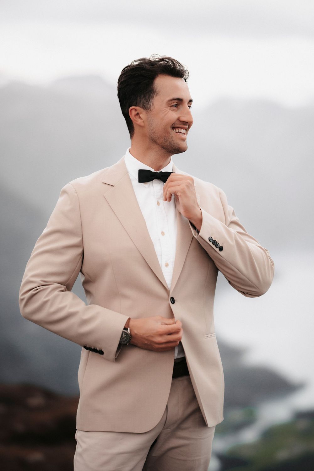 A groom in a beige suit and black bowtie adjusts his collar while smiling on a mountain overlook.