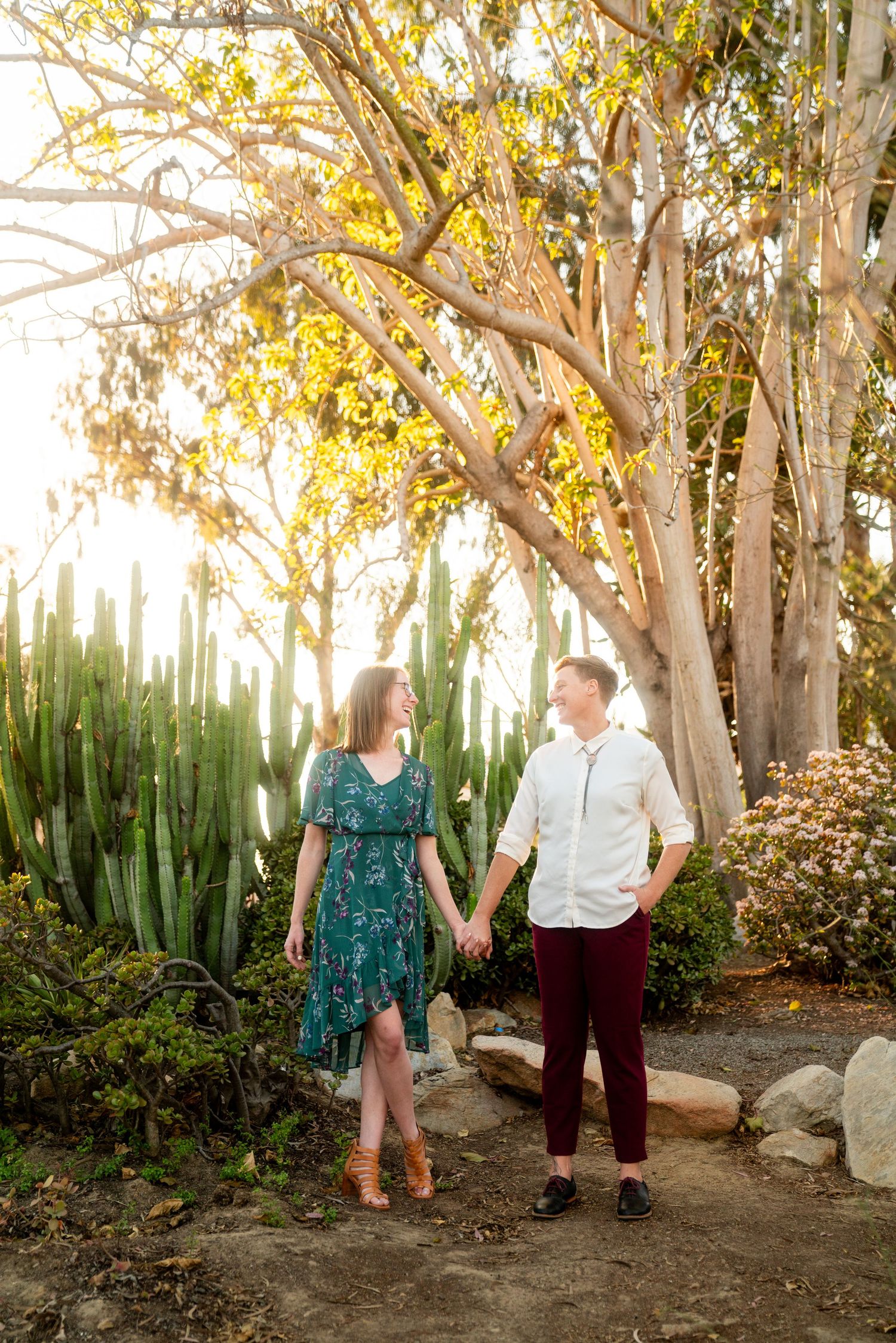 A couple holds hands while walking along a garden path surrounded by eucalyptus trees and succulents at sunset.