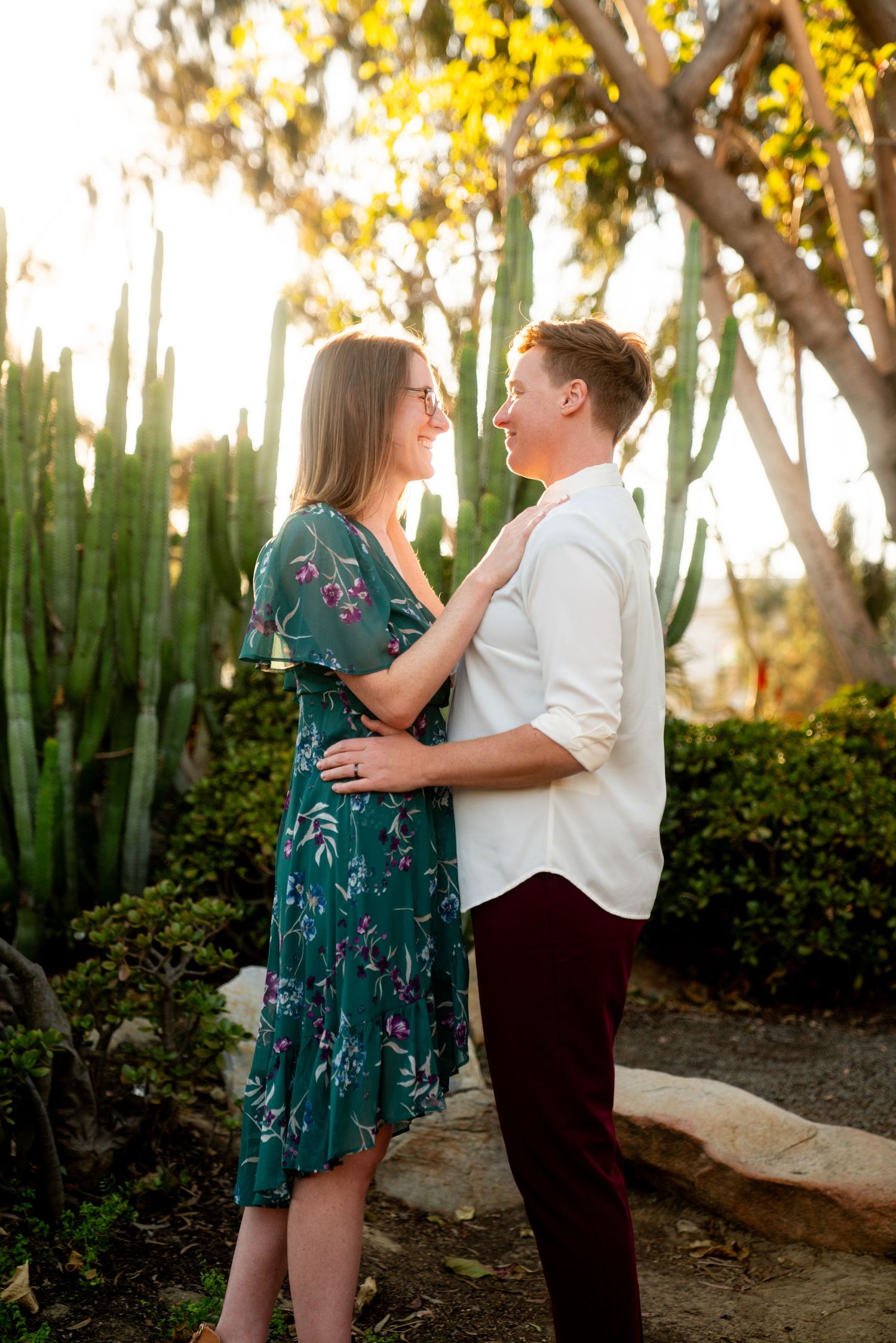A couple embraces in a romantic garden setting with yellow blooming trees and tall desert cacti at sunset.