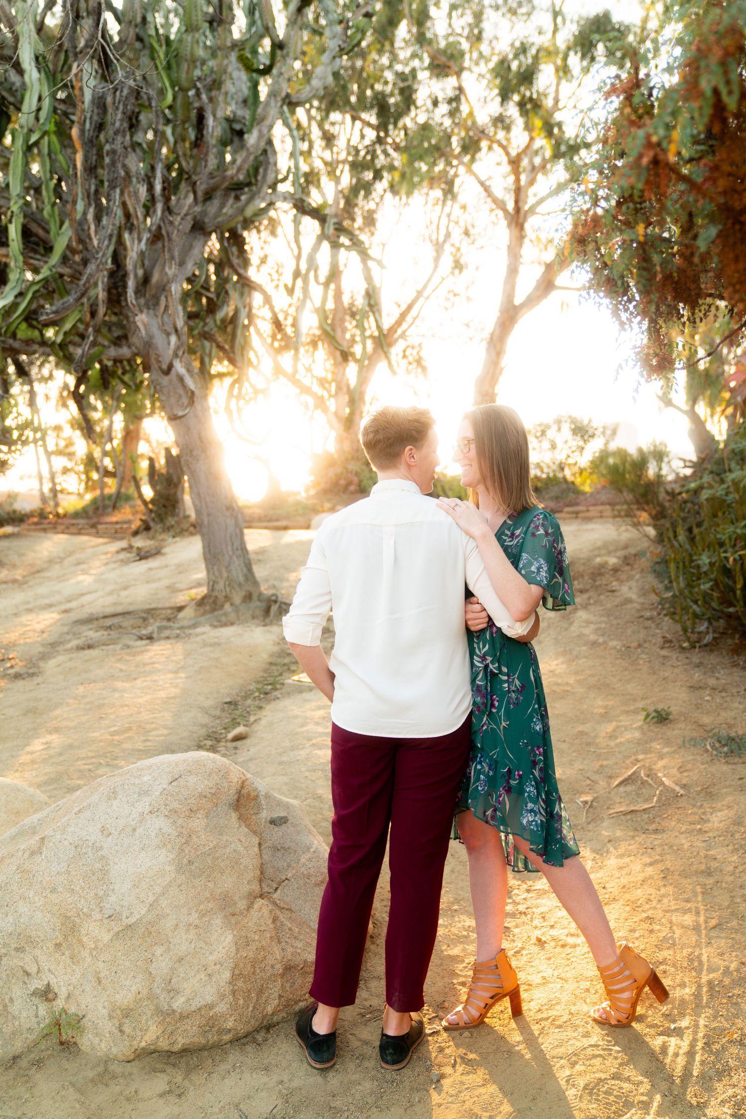 A romantic couple shares a tender moment at sunset among eucalyptus trees, she in a green dress and he in burgundy pants.