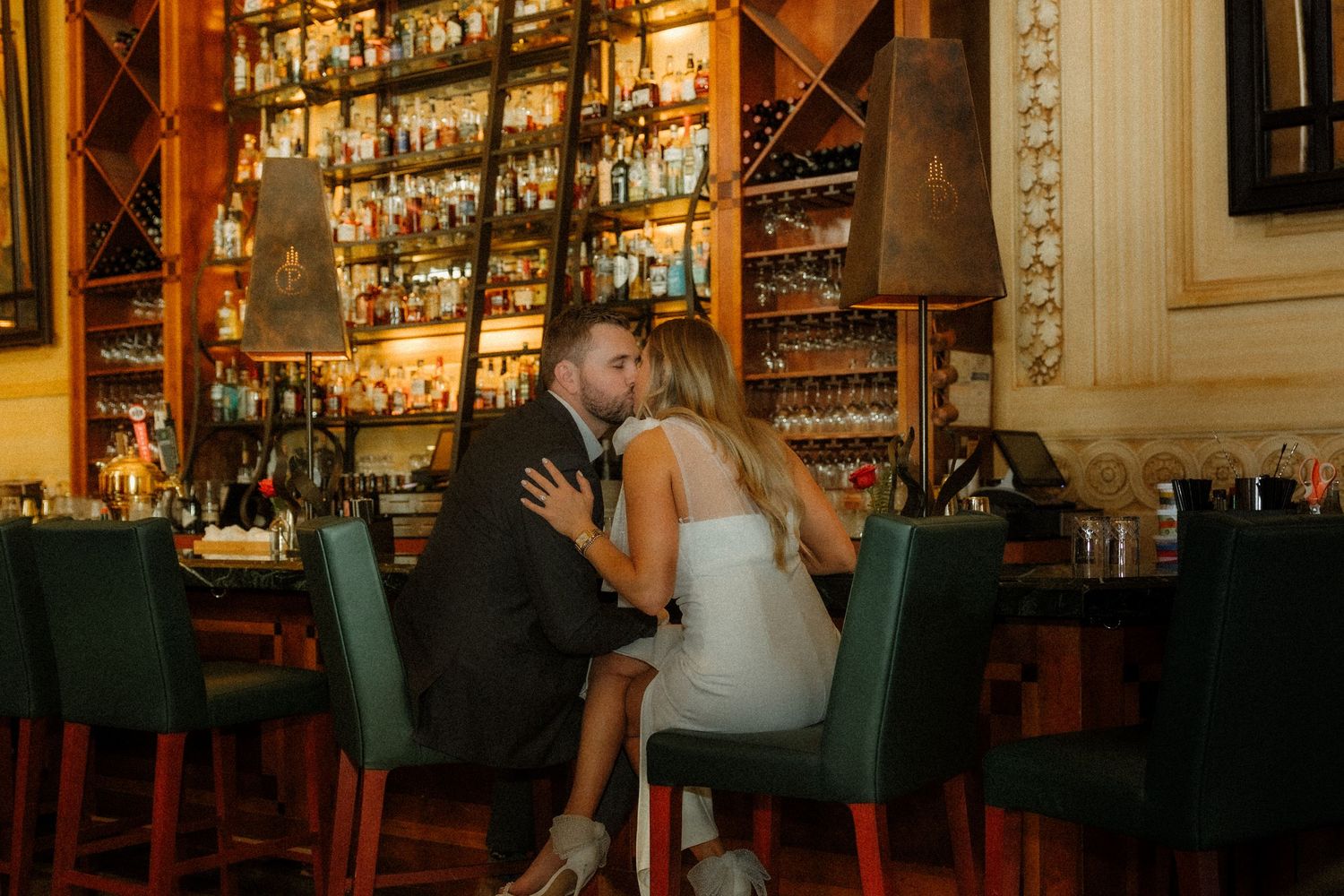A couple shares an intimate moment at an elegant bar with illuminated shelves of spirits and bottles behind them.
