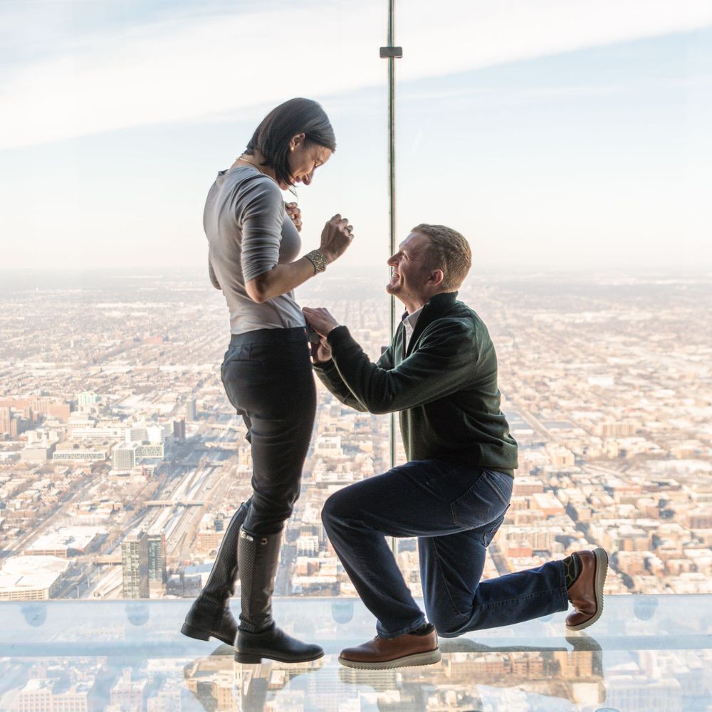A proposal moment takes place on a glass observation deck high above a sprawling cityscape below.