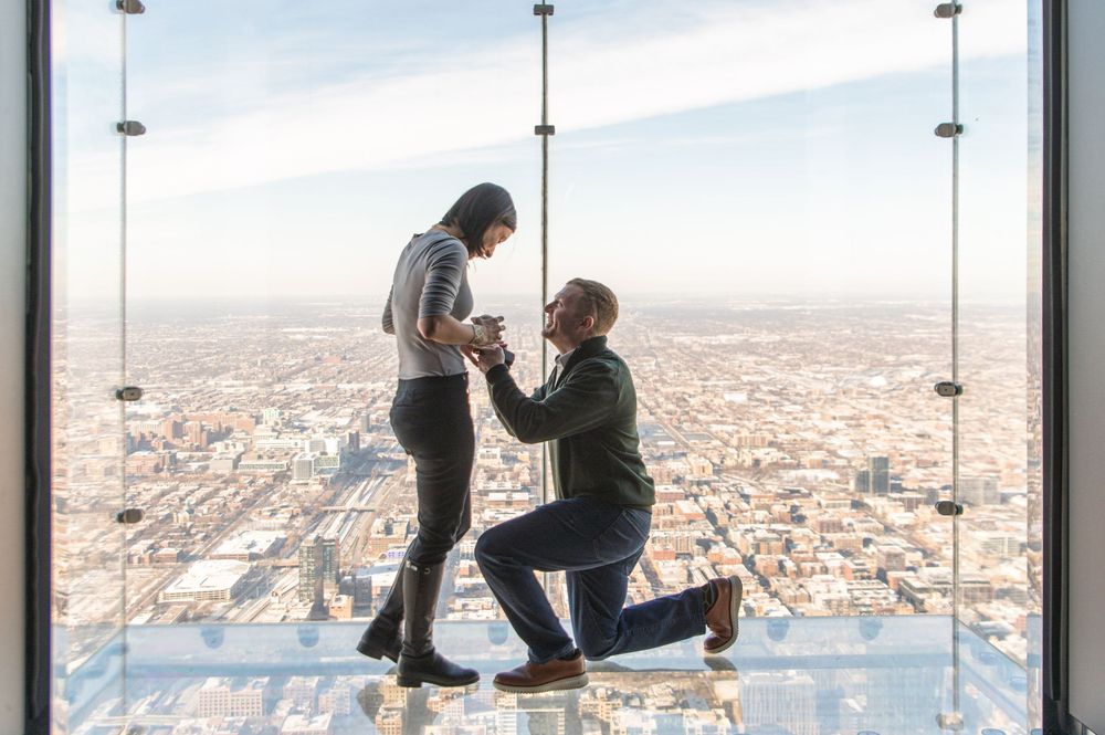 A romantic proposal takes place on the glass observation deck of a skyscraper overlooking a vast cityscape below.