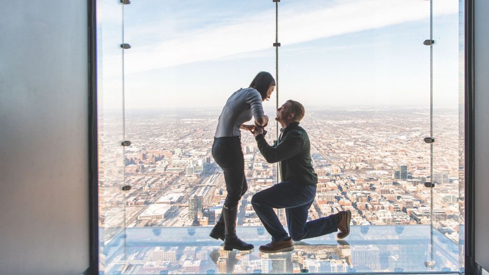 A proposal scene silhouetted against a cityscape view from inside a glass observation deck high above the city.