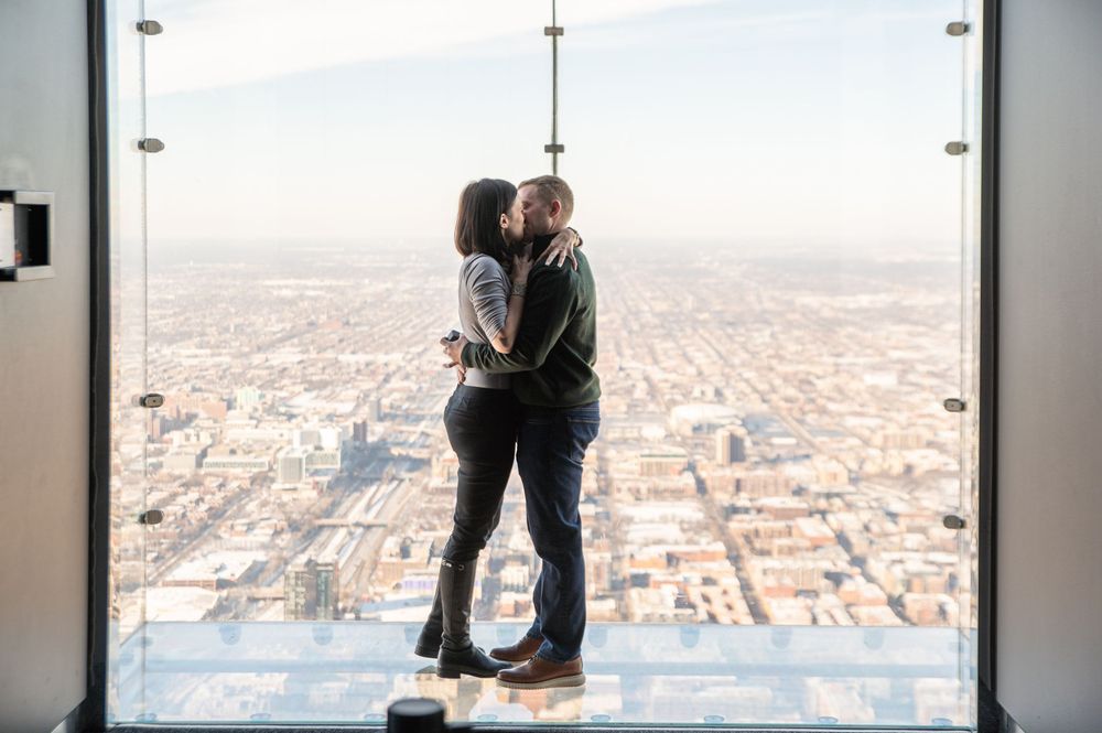 A couple embraces while standing on the glass ledge of Willis Tower Skydeck overlooking snowy Chicago cityscape.