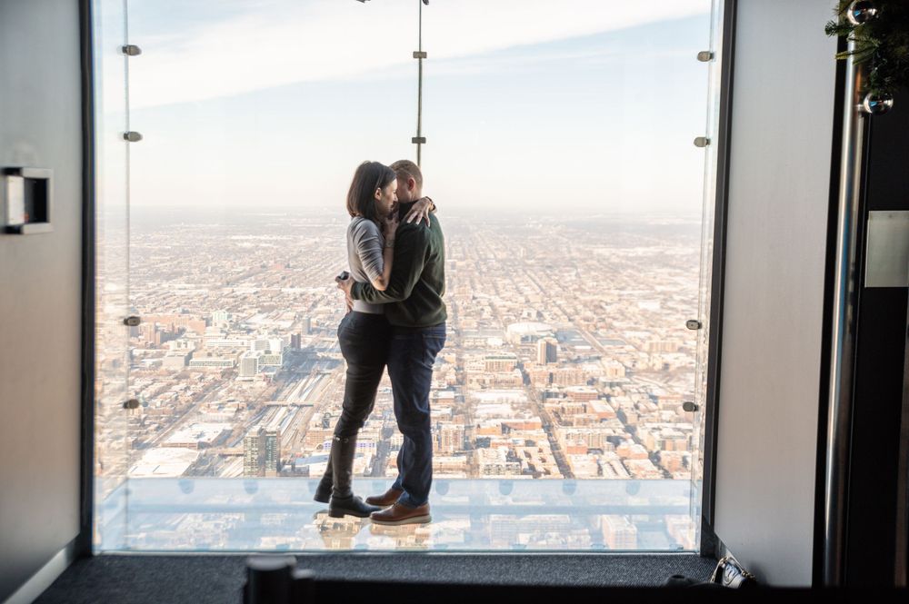 A couple embraces while standing on a glass observation deck overlooking a vast cityscape below on a clear day.