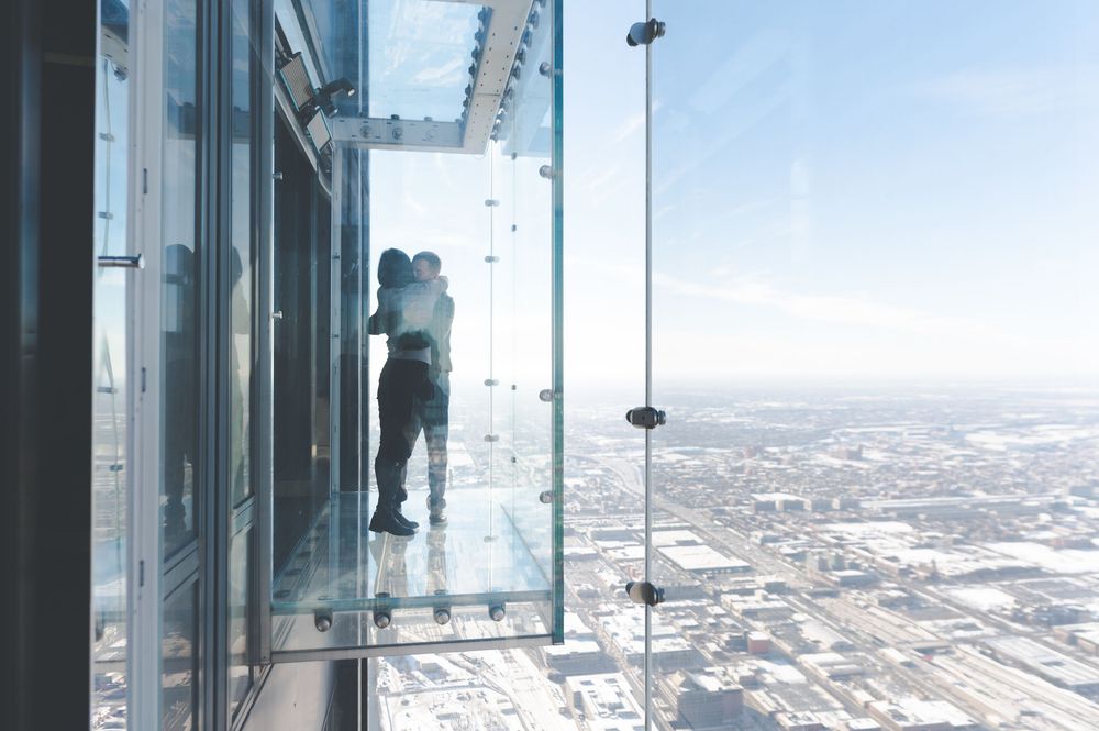 Someone stands on the glass ledge observation deck of a skyscraper with sweeping aerial views of the city below.