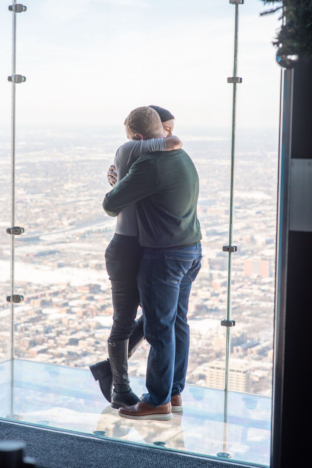 Couple embraces while standing on glass observation deck overlooking cityscape from high above.