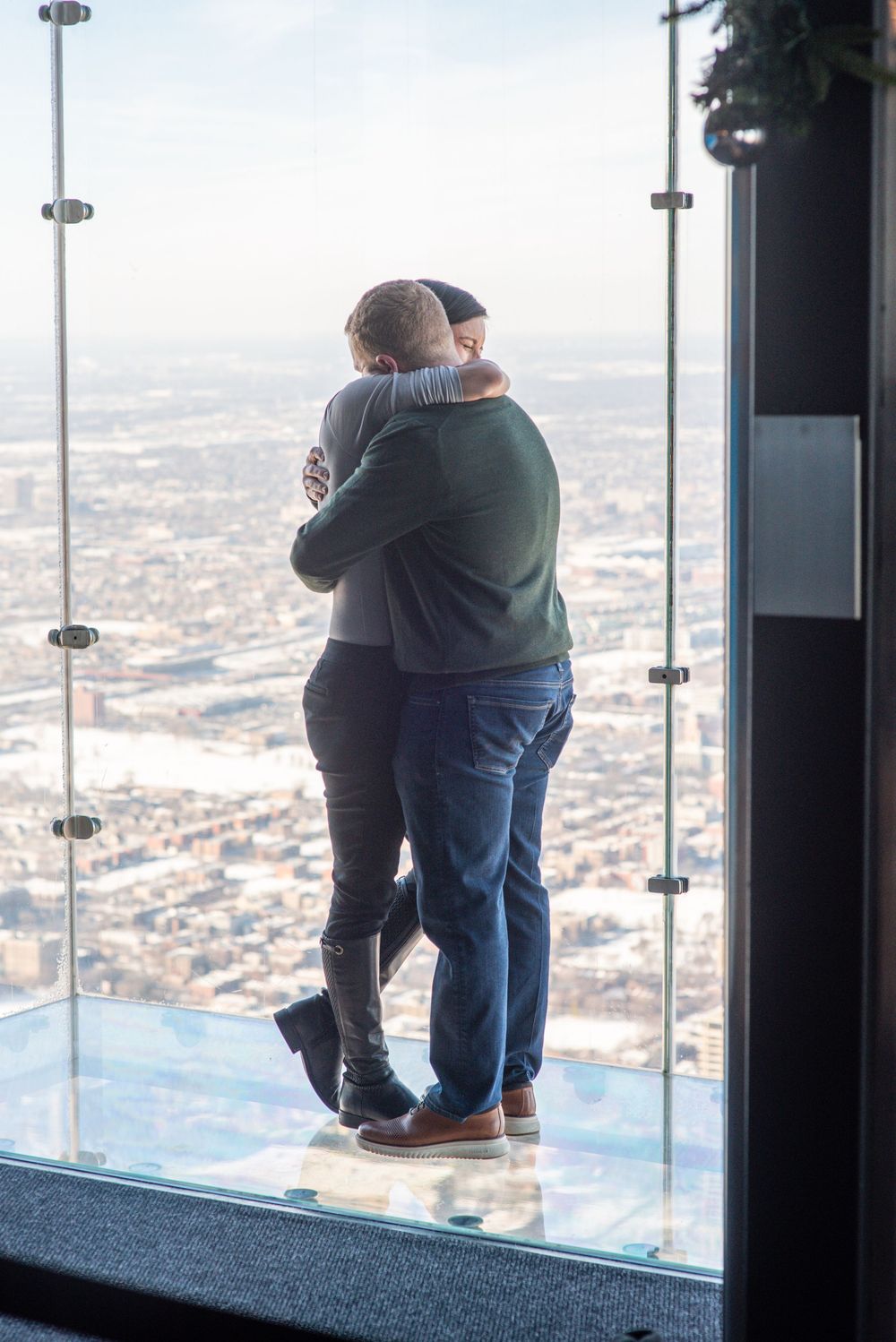 Couple embraces while standing on glass observation deck overlooking cityscape from high above.