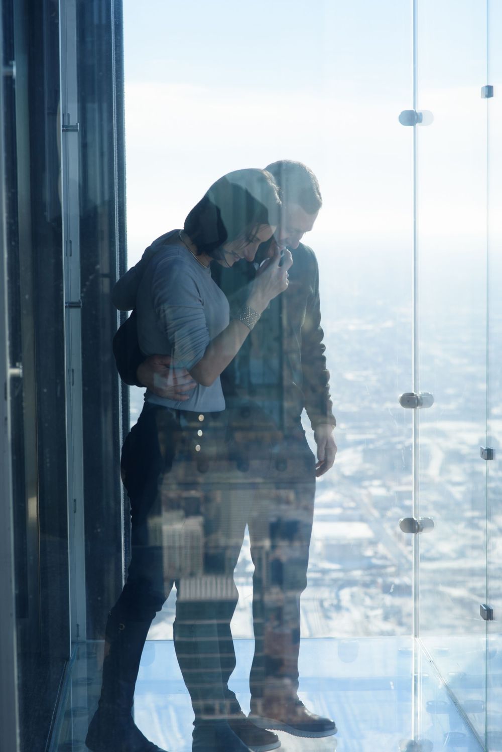 A couple embraces while standing in a glass observation deck high above a cityscape with scenic views below.