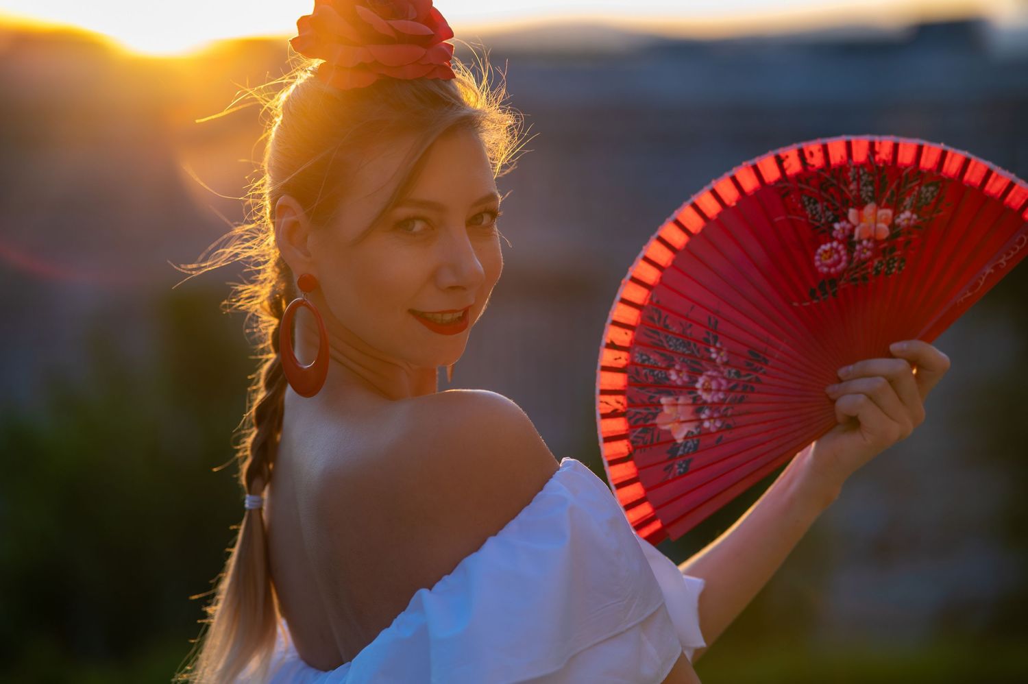 A figure in a white dress holds a red decorative fan while backlit by golden sunset light.