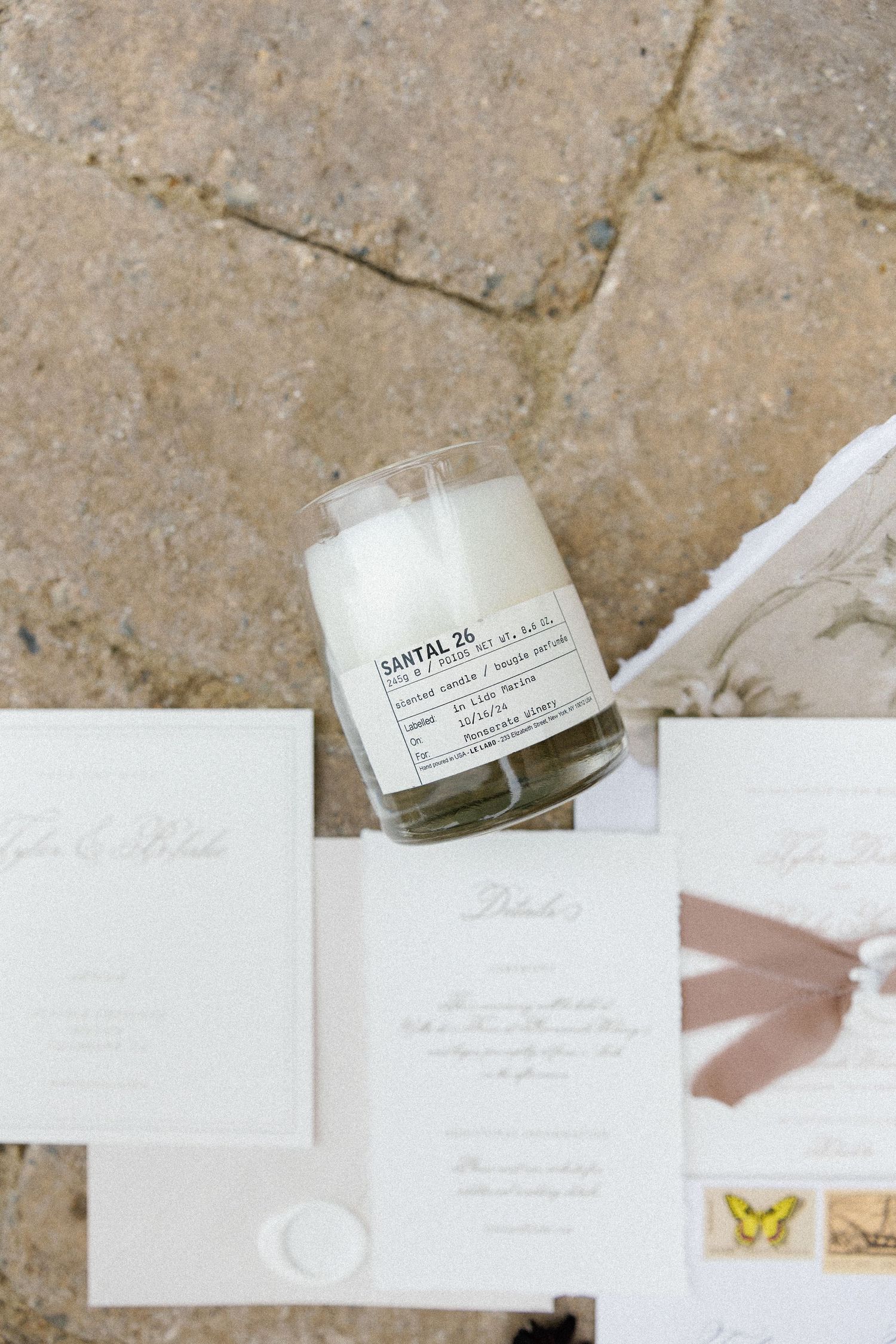 A Diptyque Santal 34 candle sits atop elegant white wedding invitation cards on a stone surface.