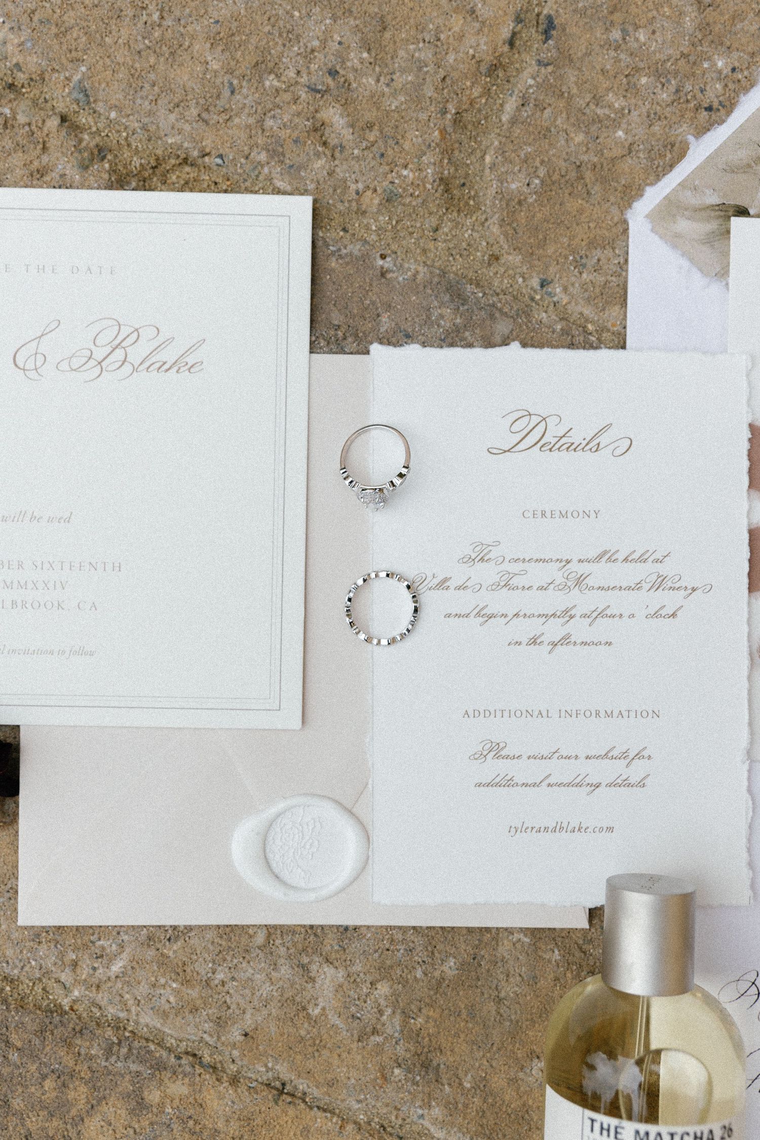 Elegant wedding invitation suite with two diamond rings and a bottle of perfume on a textured stone surface.