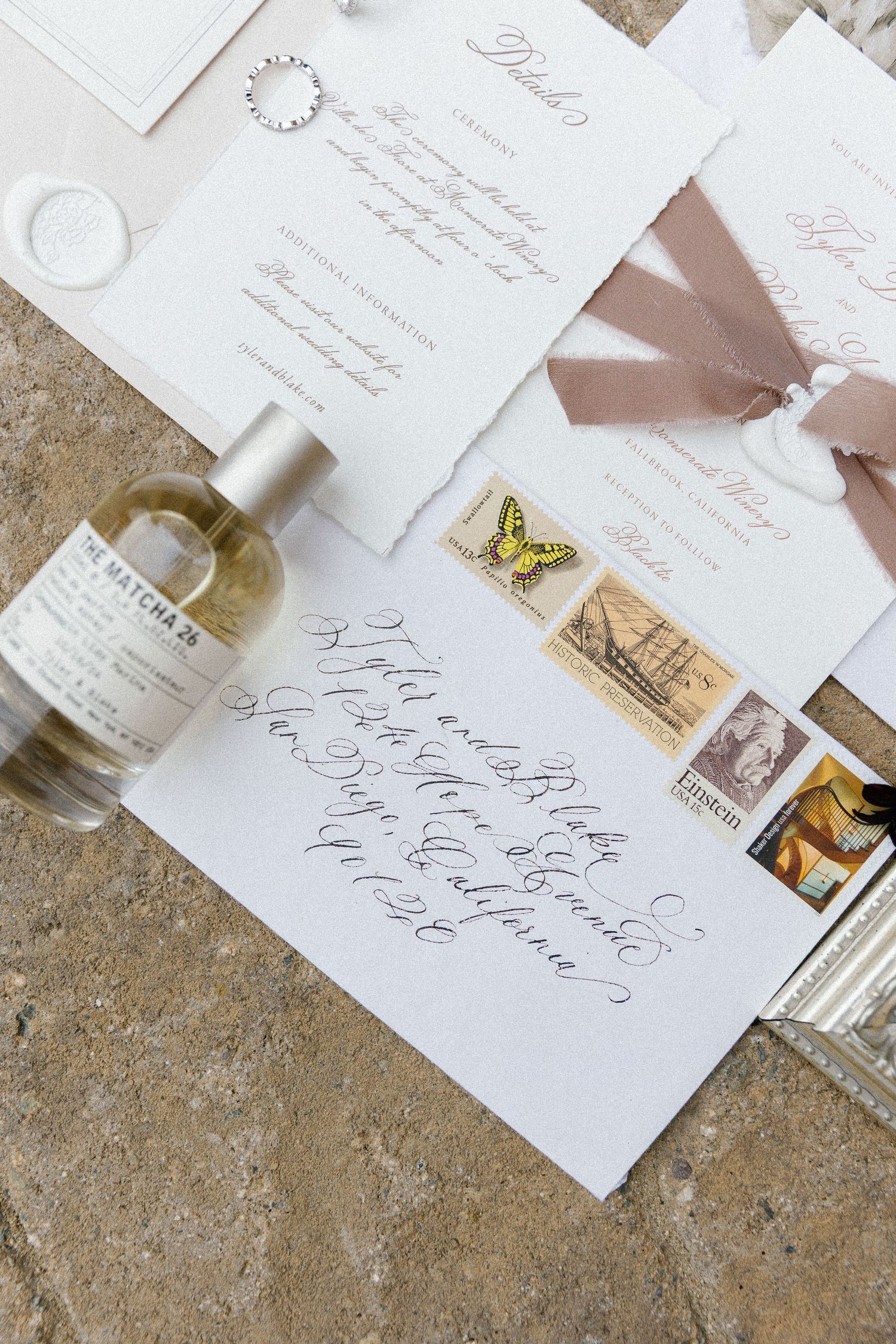 Elegant wedding invitation suite with calligraphy, vintage stamps, and perfume bottle on textured stone surface.