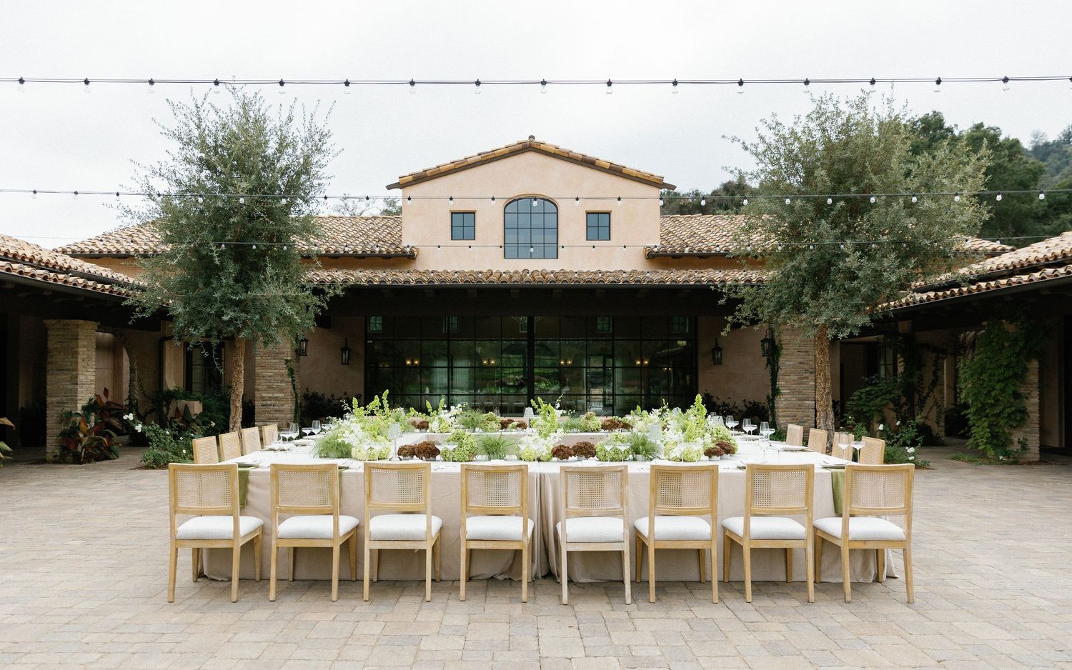 Elegant outdoor wedding reception table setup in Mediterranean courtyard with olive trees and terra cotta architecture.