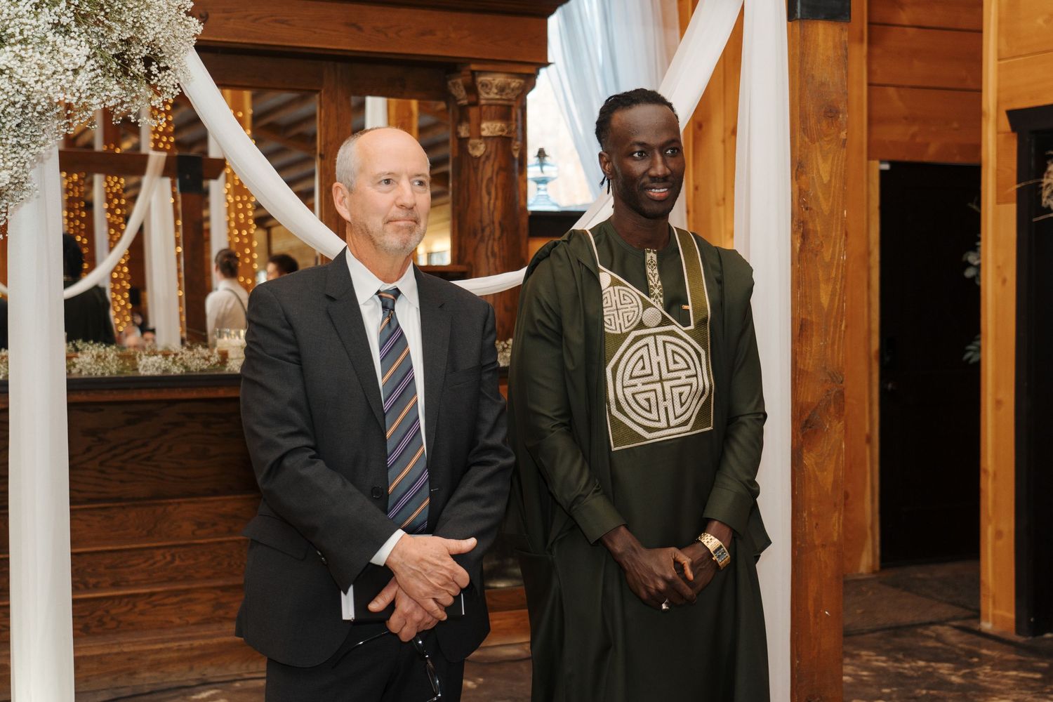 Two individuals stand together at a wooden-paneled venue decorated with white drapery and floral arrangements.