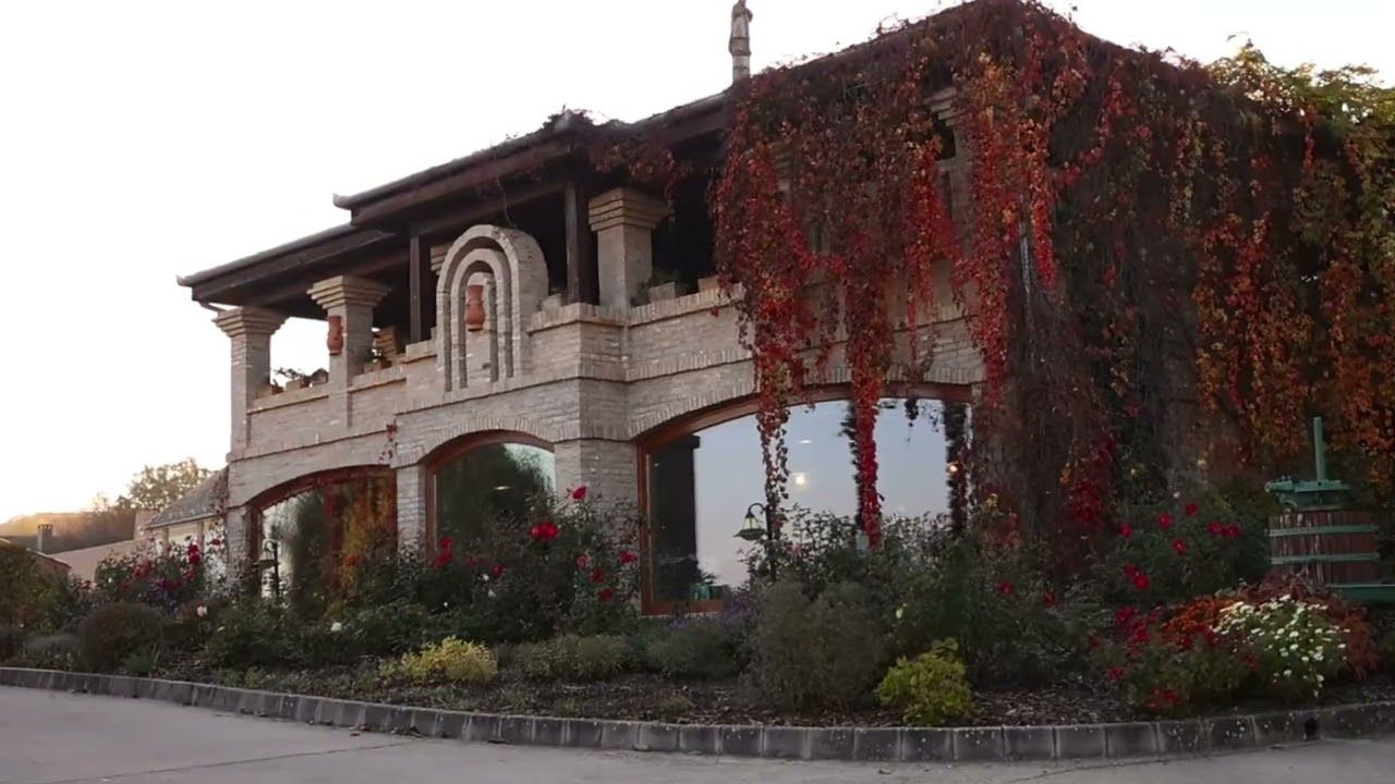 A rustic stone mansion with arched balconies and red climbing vines decorating its exterior facade at dusk.