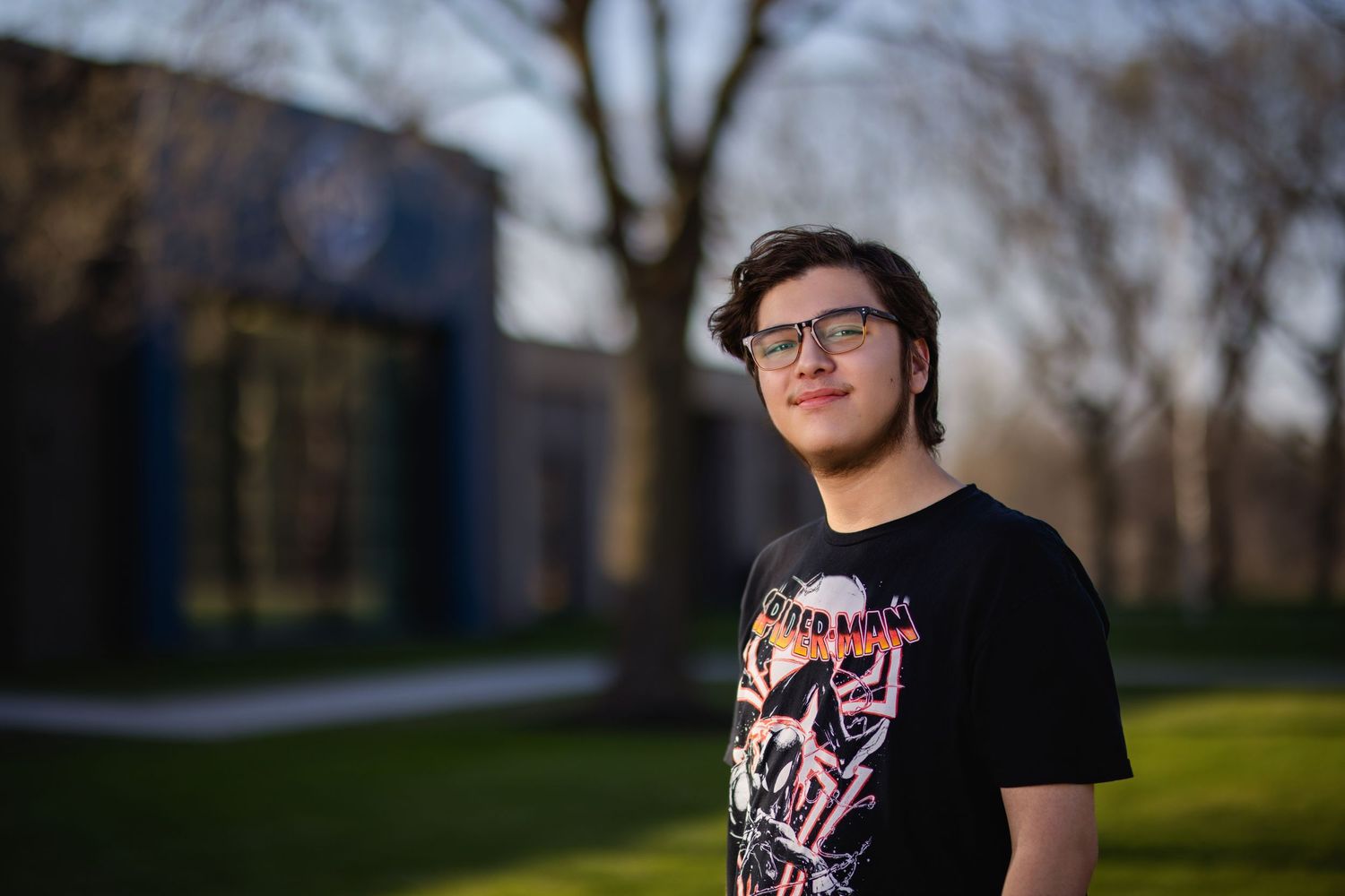 Senior wearing black graphic t-shirt stands outdoors on sunny day with trees and buildings in background. Iowa Central Community College
