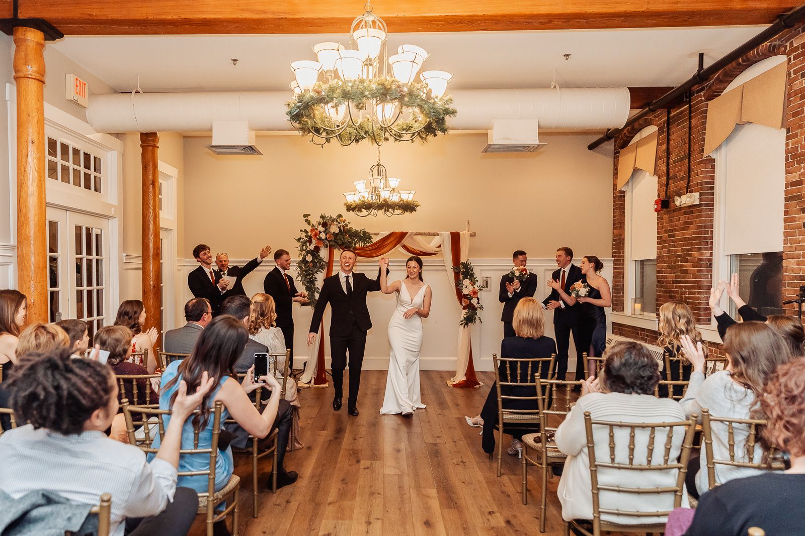 A bride and groom share their first dance under a crystal chandelier in a rustic wedding venue with exposed brick walls.