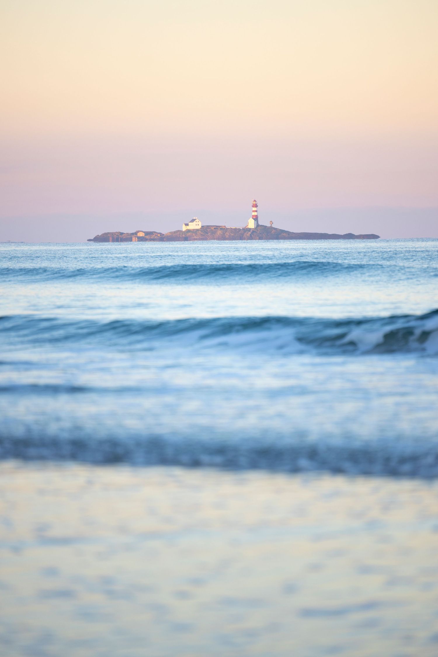 A lighthouse on a small island with waves crashing in the foreground during a pink and purple sunset.