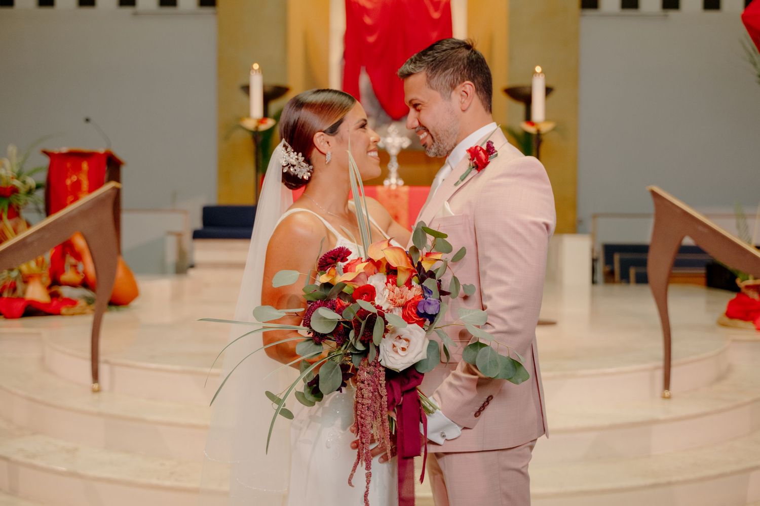 Newlywed couple stands together in a church altar holding a vibrant red and pink floral bouquet.