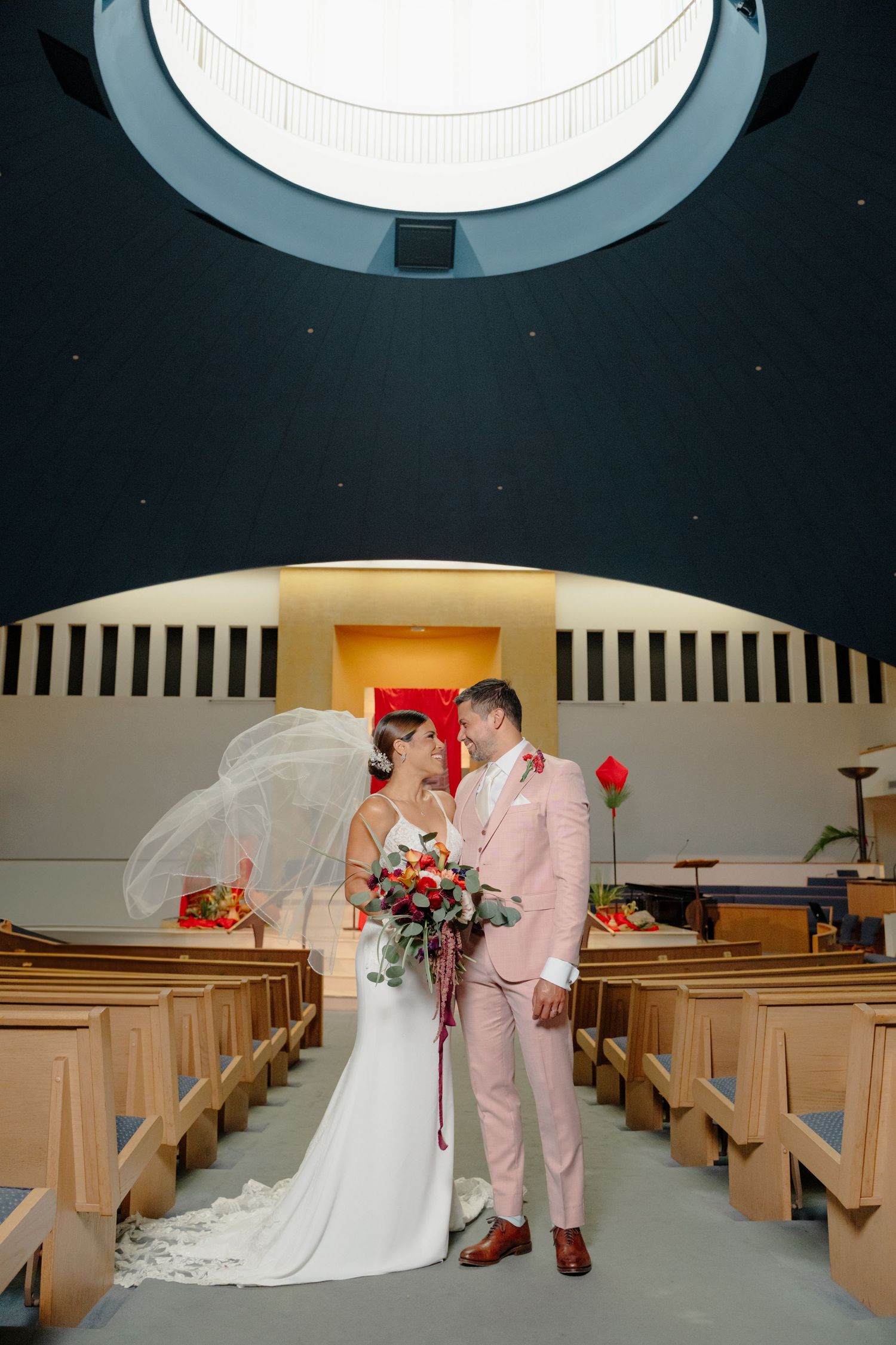A couple shares an intimate moment in a church aisle with white and pink wedding attire while holding red roses.
