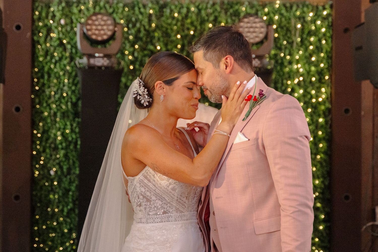 A romantic wedding moment with the bride in a white gown and groom in pink suit embracing against a leafy green backdrop.