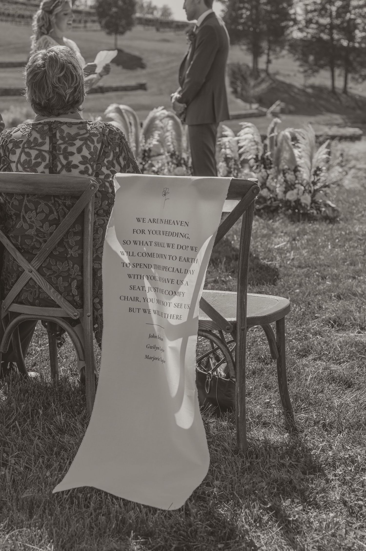 A chair with a memorial message on white sign displayed at an outdoor wedding ceremony setting in black and white. by Samantha campbell