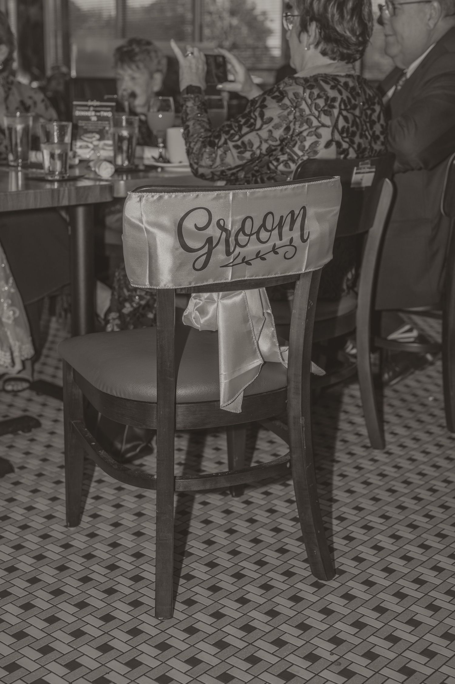 A black and white photo by Samantha Campbell Imagery of a wooden chair with a 'Groom' sign attached to the back at a wedding reception.