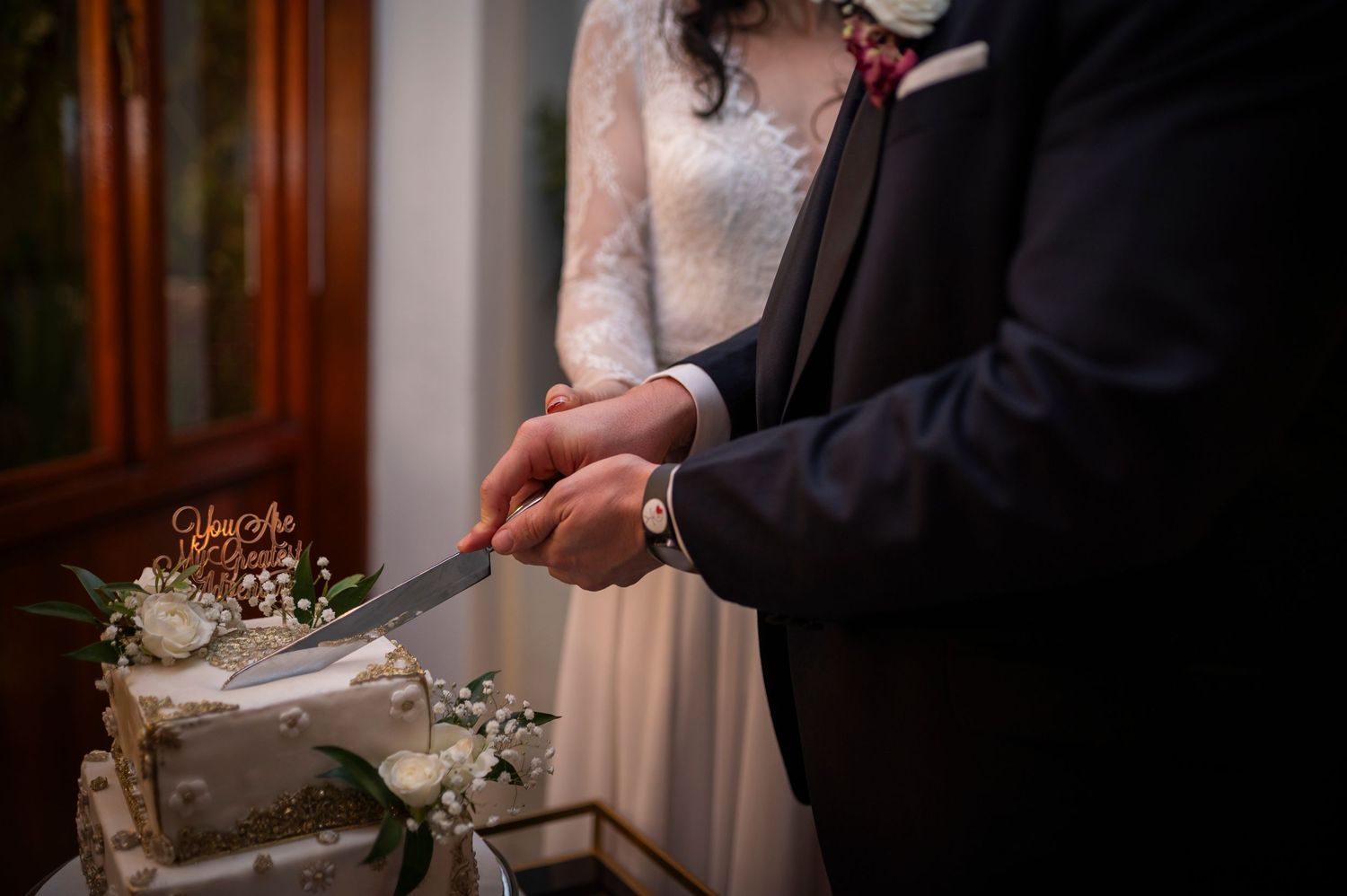 Newlyweds cut their white floral decorated wedding cake together with hands intertwined on the knife.