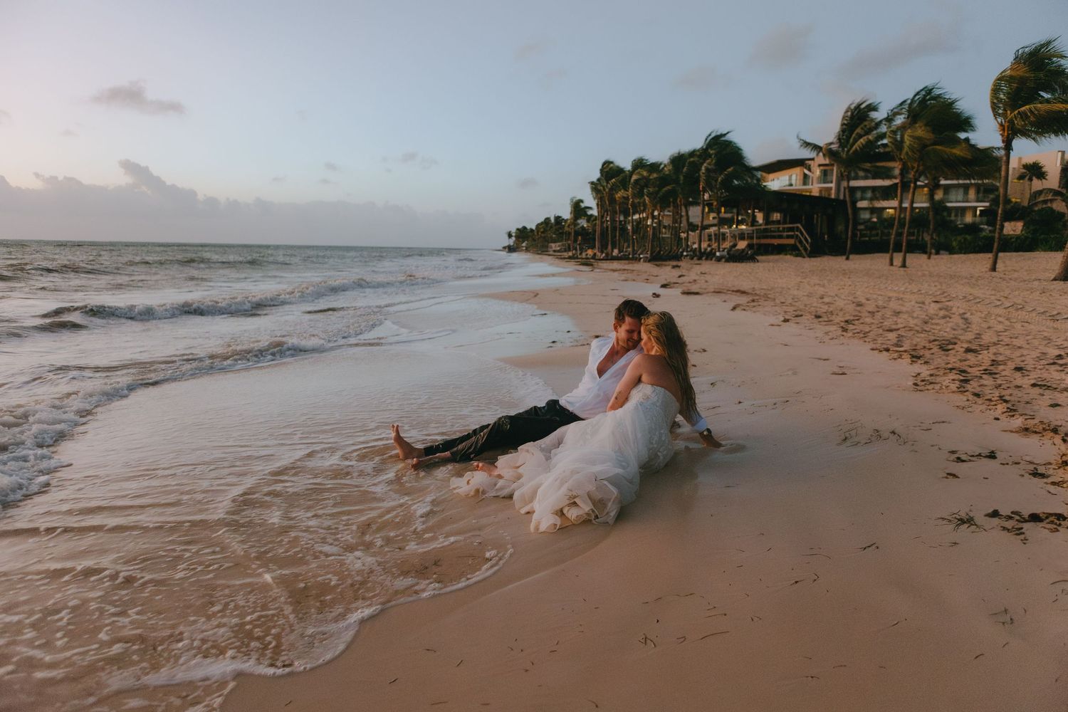 A couple embrace joyfully on a beach, with the woman in a white dress being lifted up against a cloudy ocean backdrop.