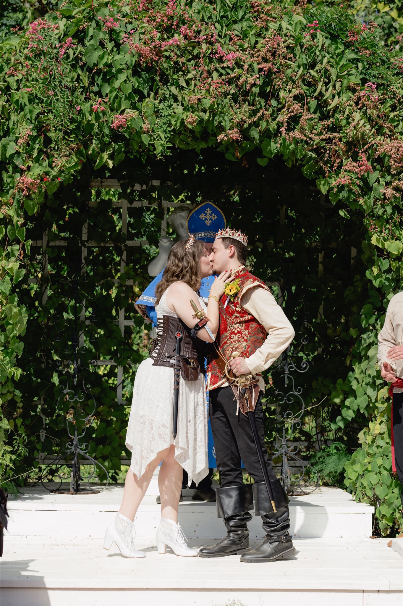 Medieval costumed couple shares a romantic moment in a garden setting with flowering vines and white walls.