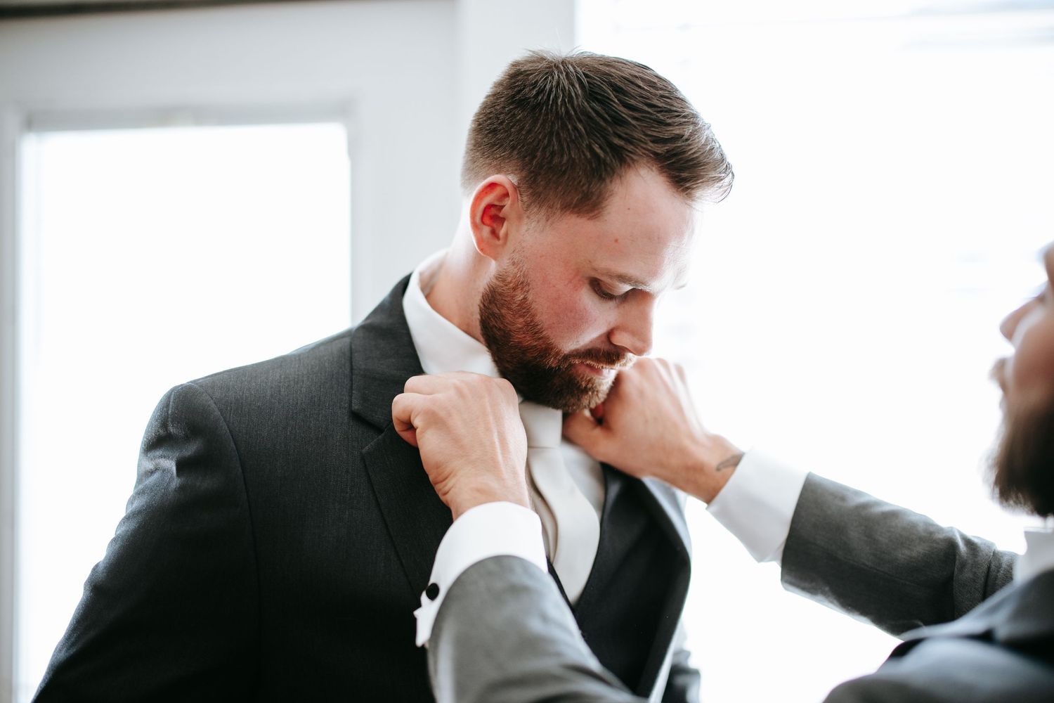 Someone adjusts the bowtie on a groom wearing a dark suit during wedding preparations.