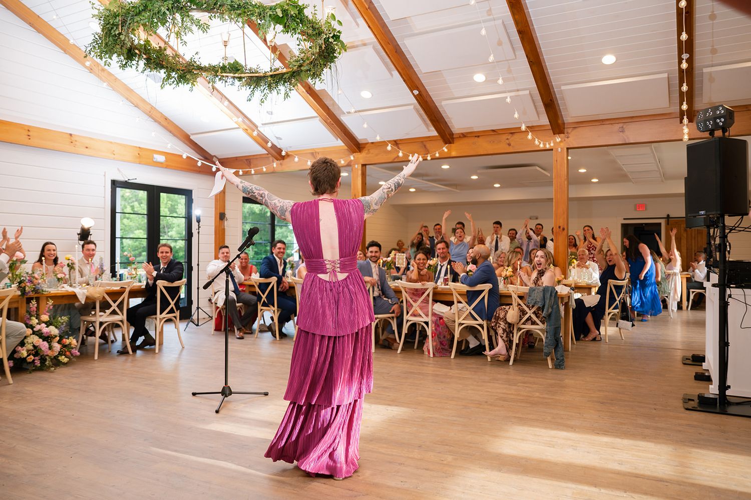 Someone in a floor-length magenta dress performs in front of an audience in a bright rustic event venue.
